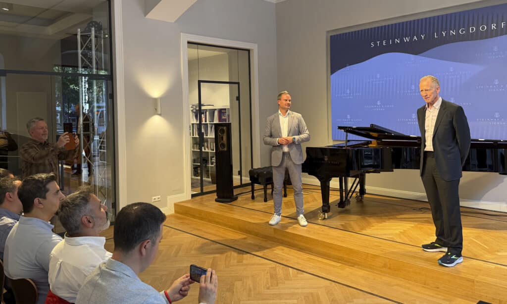 Peter Lyngdorf presenting at a Steinway Lyngdorf and Steinway & Sons event in Vienna, standing beside a Steinway grand piano in front of an audience.