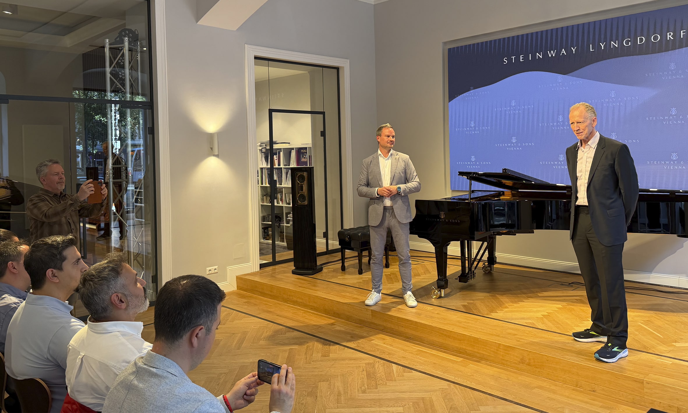 Peter Lyngdorf presenting at a Steinway Lyngdorf and Steinway & Sons event in Vienna, standing beside a Steinway grand piano in front of an audience.